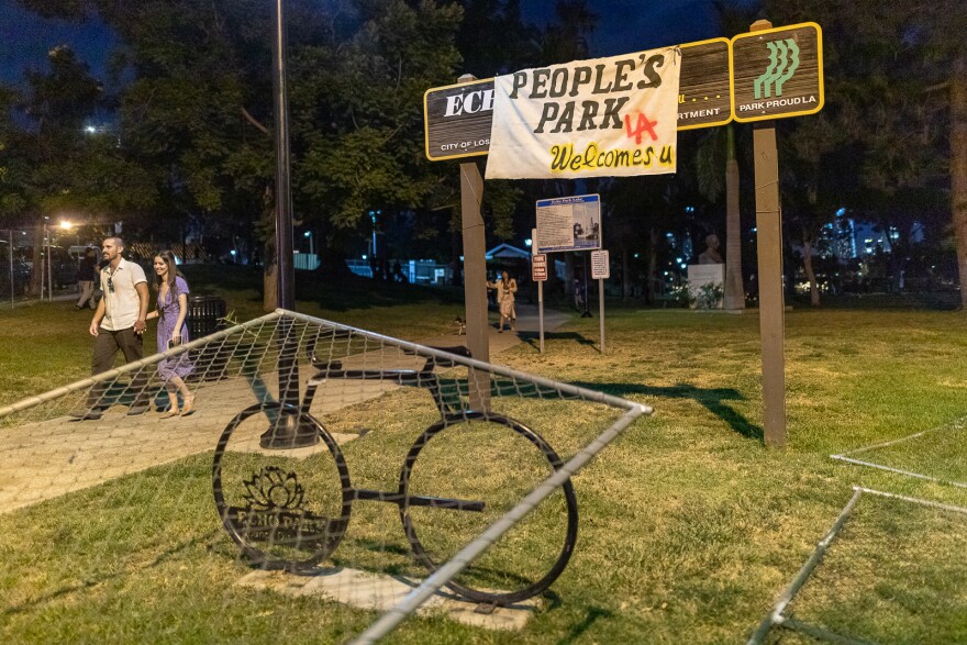 Fencing rests on the ground and on top of a bike sculpture at Echo Park. People walk along a nearby path. A whiet banner reads "PEOPLE'S PARK LA Welcomes U," draped over the park's own official sign.