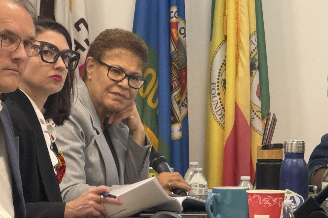 A woman with medium skin tone and short curly hair wearing black rimmed glasses and a gray blazer sits at a table with her head resting on her hand. Behind her are various flags. 