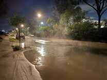 The sewage spill floods the street and sidewalk. The sewage is brown and cloudy against a street lamp light.