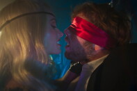 Olivia Wilde wears a blonde wig and eyepatch, face directly in front of Cooper Hoffman in a suit and red blindfold.