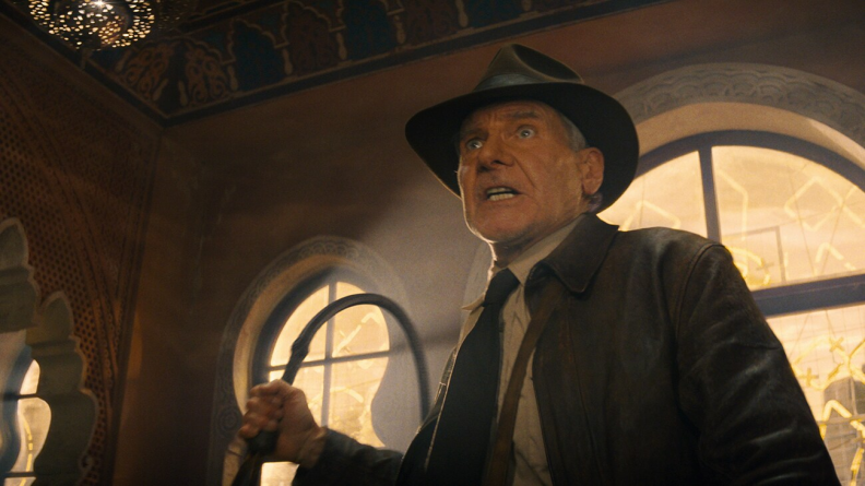 Harrison Ford stands in a large room that seems to be inside a temple or church of some sort. The walls are made of sand colored stone with large windows of curved shapes. The windows let warm light in from the outside. Ford wears the classic Indiana Jones outfit: a brown hat, brown leather jacket, whit button-up and black tie. He raises his iconic whip in his right hand, bracing to use it. His face looks like one of shock or surprise, as if caught off guard by whoever stands in front of him out of view. His eyes are wide, his mouth slightly open but tensed. 