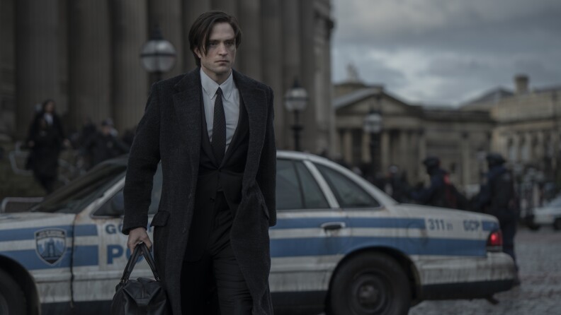 A man in suit and coat carries a bag in one hand. He has a grim or determined look on his face and is walking away from officers and a grimy police squad car.