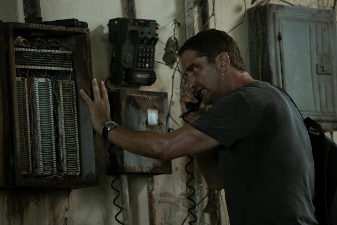 Gerard Butler stands in a dilapidated room. The white walls are stained with brown rust, a communications radio and wall phone hanging on the wall, warped and crusted with age. Butler holds the wall phone to his ear, only his left side visible as he braces himself against the wall with his left hand on the communications radio. He wears a dark gray shirt that has sweat stains. His faces is smudged with dirt and shines with sweat. His faces looks panicked and out of breath. 