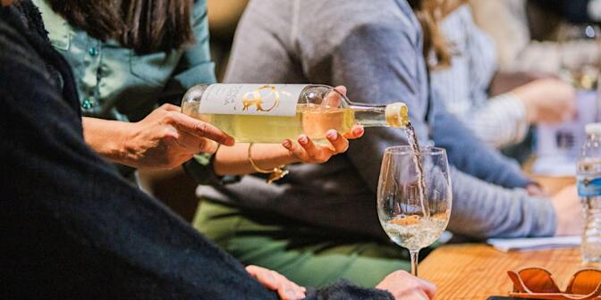 A pair of hands pours a glass of white wine as people sit a table, with one person holding the glass still. 