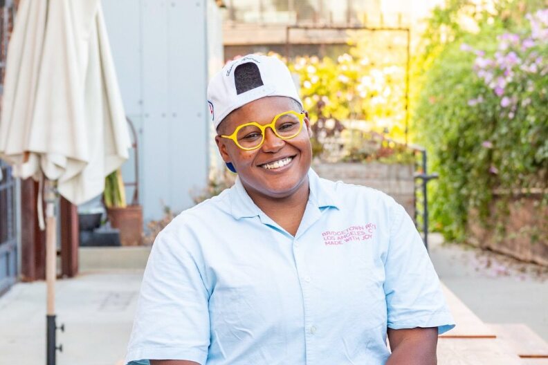 Chef Rashida Holmes, a dark-skinned woman, stands in front of a picnic table in a blue button-down shirt and yellow-framed glasses, smiling at the camera.