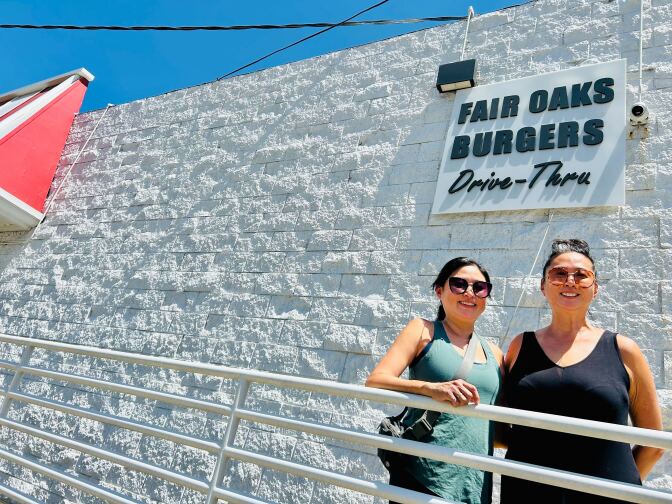 Two women stand in front of a white wall. A sign above them reads "Fair Oaks Burgers, Drive-Thru."
