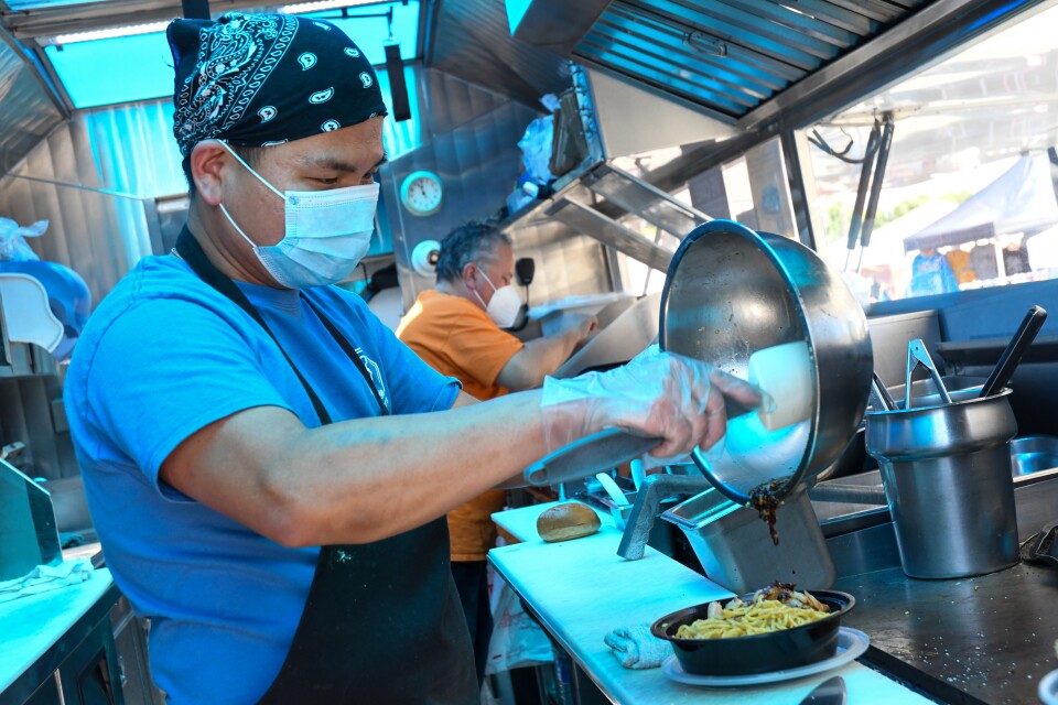 A masked man prepares food for customers as takes orders at a food truck.