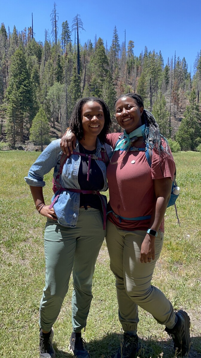 Two Black women, clad in hiking gear, hug each other and smile. There is a forest in the background. 