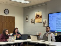 Three people sit in a small classroom, with their laptops in front of them. One of the people --- a man with medium skin tone, short dark hair, a long-sleeved shirt, and black-framed glasses-- gestures while explaining something.  Behind him, a document containing the words "Circumstances of Death" is projected on a large screen. 