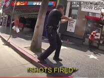 A screen shot from the officer body cam footage of the fatal shooting of Matthew Sova in 2021 shows officers pointing their weapons at Sova, who wears a red shirt and dark pants and is standing in front of a business in Hollywood. 