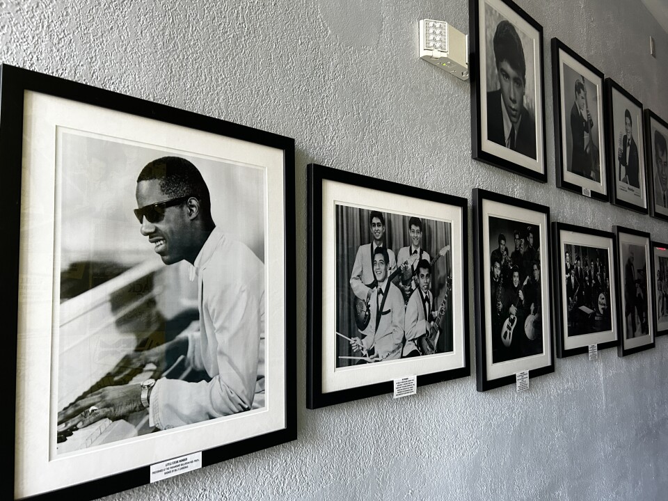 Black and white photos of artists including Stevie Wonder and Little Ray Jimenez line a white wall. 