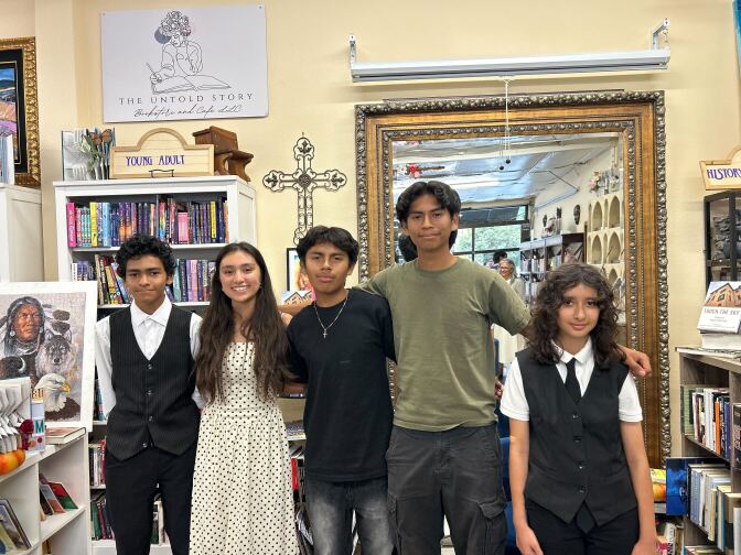 A group of students stand in a bookstore with their arms around one another. In the background, a small gray sign reads, "The Untold Story." 