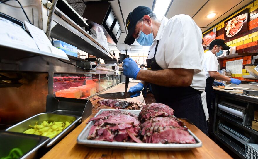 A restaurant kitchen employee wearing gloves and a mask uses a knife to slice meat on a wooden counter.