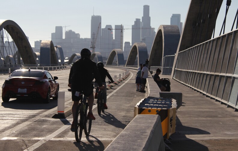 Two people ride bikes in a bike lane on a bridge as a red car drives past them on the left. A group of people are standing in the bike lane further up the road. The downtown Los Angeles skyline is visible in the distance.