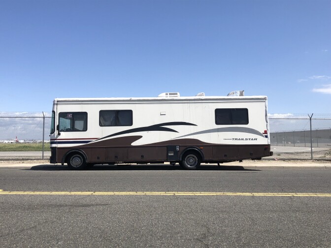 An RV is parked along a street next to a fence topped in barbed wire.