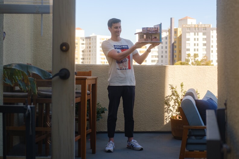 A view of Wright standing on his balcony outside holding the Al & Bea's miniature against the skyline. He's smiling and looking at it.