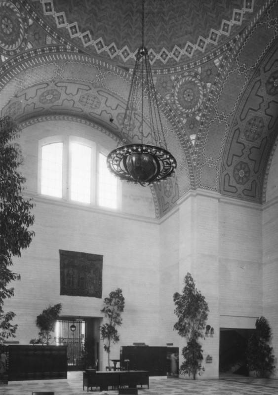A black and white archival view of the ornate mosaic-like dome of the library's rotunda. The sunburst image located directly above the globe chandelier mirrors the sunburst design of the pyramid on top of the building. A chandelier hangs from the rotunda's ceiling, composed of cast bronze, is part of a model of the solar system.