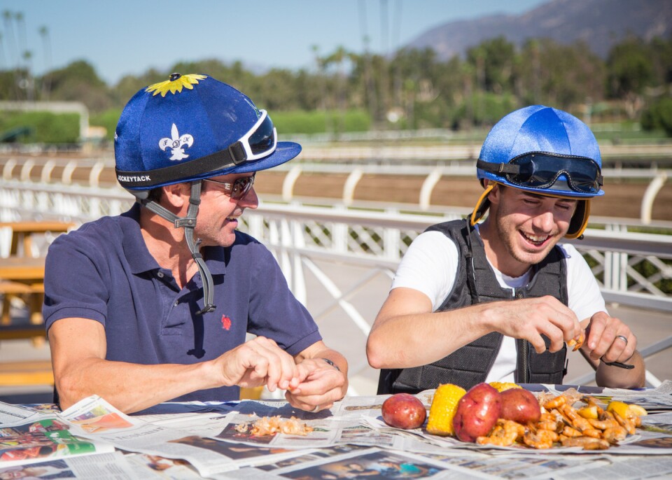 Two jockeys eat a shrimp boil trackside at Santa Anita Park. The shrimp, corn and potatoes are atop of newspapers.