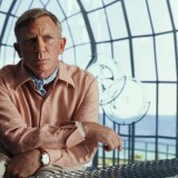Daniel Craig sits at a grey wire table. His right arm lays on the table as his left arm lays crossed over on top of it. Craig leans slightly forward as he looks at something out of frame. His brows are slightly furrowed with lips pursed as if puzzled. Craig wears a peach colored button-up. A light blue ascot can be seen peeking out from the open collar of the shirt. The room he sits in seems to be made entirely of glass windows, the wire frames creating a grid pattern with the vast mountain landscape outside. There are various clear, glass-blown figurines around the room as well.