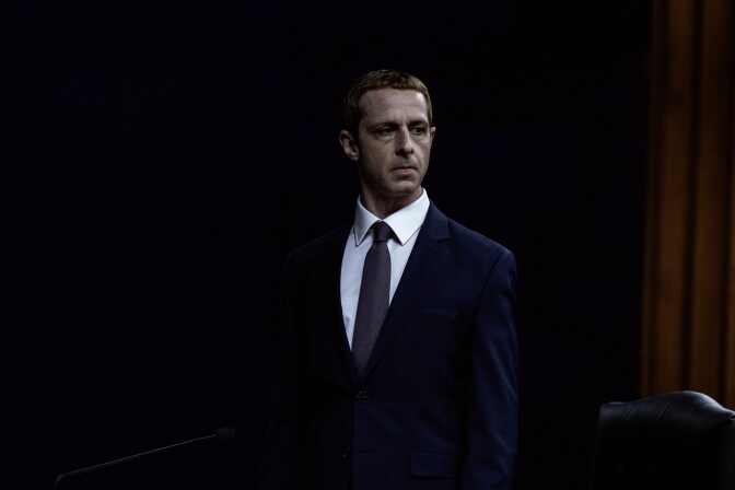 A male-presenting person with a strawberry blonde buzz-cut stands in a dark room wearing a white button-up dress shirt, a gray tie, and a navy blue suit jacket. There is a yellow curtain behind him on his right, and the upper part of a leather chair is visible in the bottom right corner of the image.