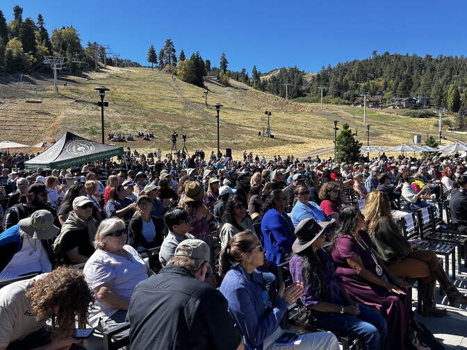 A seated crowd gathers outdoors on a ski slope under clear skies, with chairlifts and pine trees in the background.