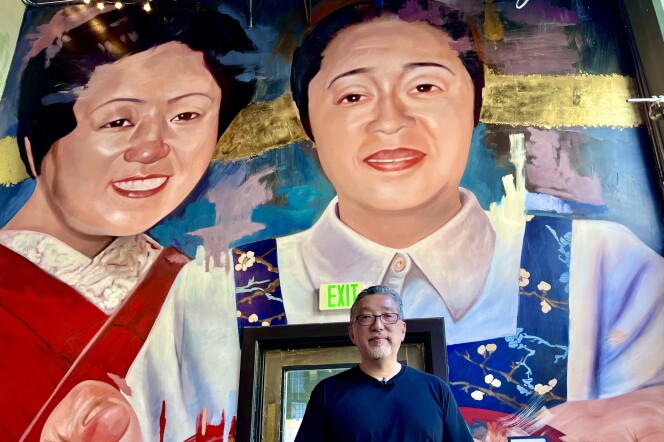 A 61-year-old Japanese American with glasses and a gray goatee stands in front of an indoors mural of two smiling Japanese American women. 