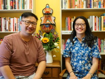 Two people with glasses stand in front of a few bookshelves lined up against a yellow wall.