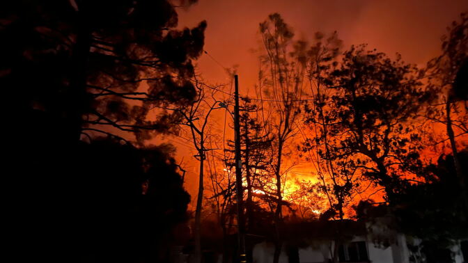 The yellow, orange, and red glow from a fire behind a home and trees at night 