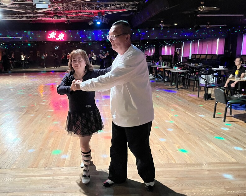 An Asian American woman in a black shirt and flowy mini-skirt dances in a ballroom with an Asian American man in a long-sleeved white shirt and black pants.