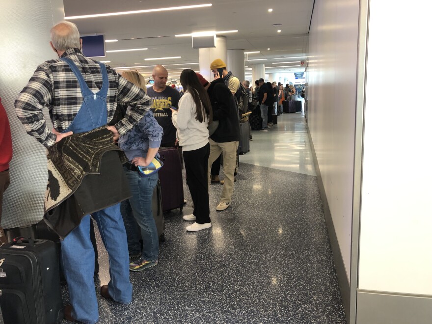 A long line of people with luggage in an airport with white pillars and walls and a speckled shiny floor.