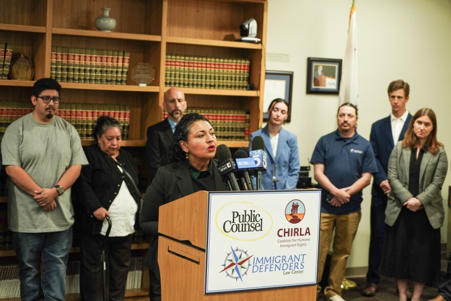 A woman speaks into microphones at a podium, with several people standing behind her.