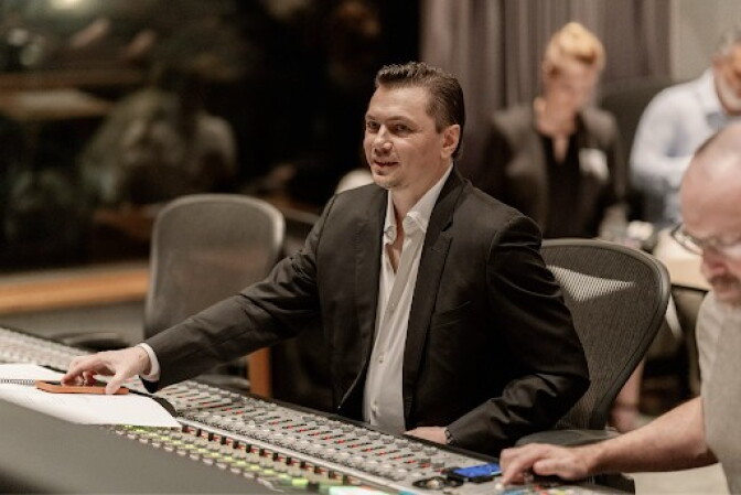 A white man with slicked back hair in a suit jacket. He is seated behind a mixing board.