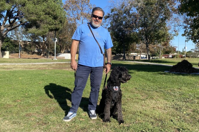 Eddie Aldrete stands on green grass at a park, holding the lede of his dog. The dog is a black Australian Labradoodle named Dakota. 