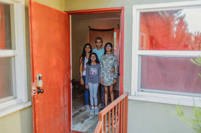 Four people stand in the doorway of an apartment with a faced dark orange door. The father, in the middle back, has his hands around the mother's waist on his right and a daughter on the left.