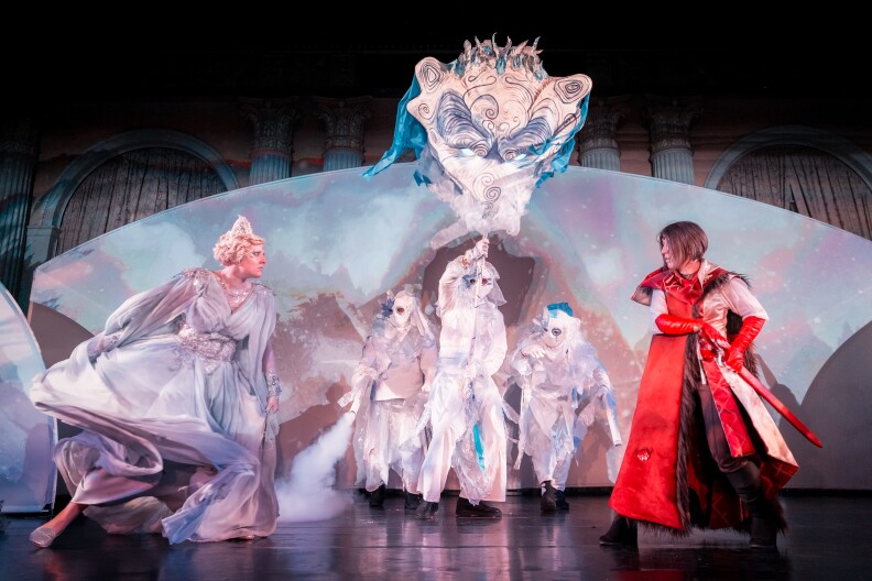 A person in a red cape and carrying a red sword prepares to face off against another person in a cloud-like ball gown and tiara.Other masked figures in white carry the figure of a creature on a pole.
