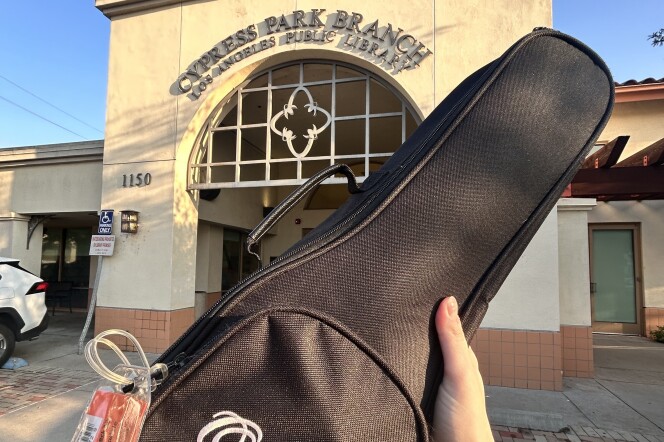 A hand is holding up a black case that says "Ohana Ukuleles" in white font on the front. A large orange laminated tag can be seen on the side, and a tan and brown building is in the background. The building has the text "Cypress Park Branch Los Angeles Public Library" above the entry. 