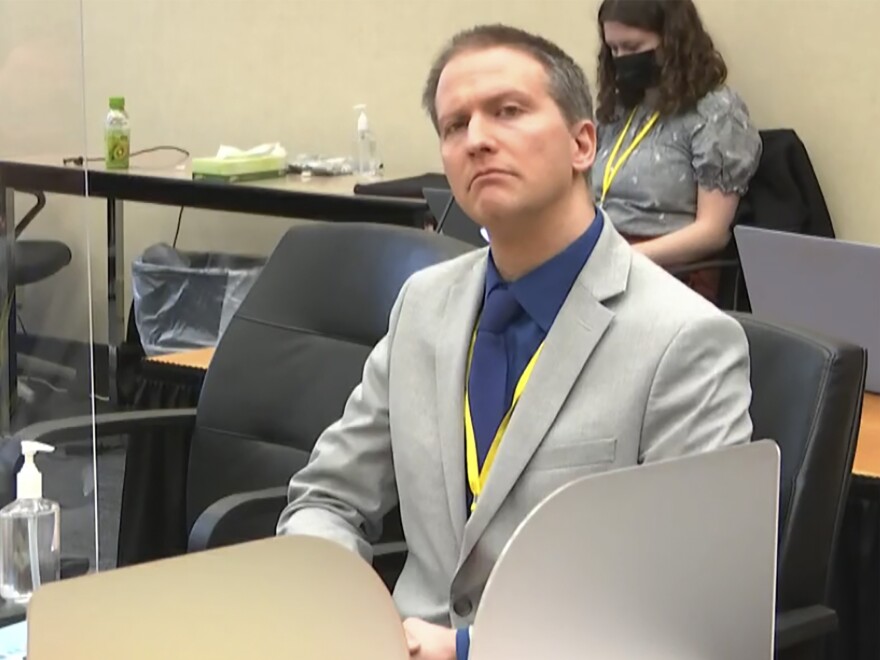 Former police officer Derek Chauvin is shown seated in a Minneapolis courtroom, wearing a light gray suit with a dark blue shirt and dark blue tie and a yellow lanyard around his neck, looking up toward the bench.
