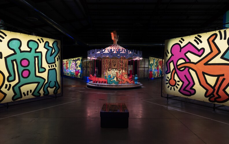 A series of panels containing Keith Haring's distinctive line drawings in pink, orange, and teal flank a carousel with similarly-shaped seats