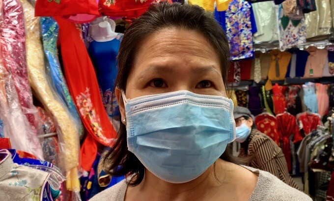 A woman of Asian descent in a mask faces the camera in a clothing stall.