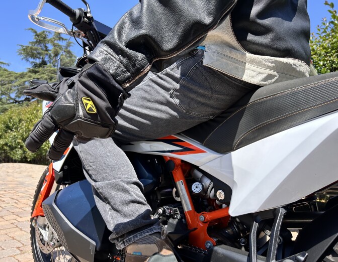 A close-up of a man on a blue, white, and orange motorcycle from the waist down. He's wearing brown boots that are resting on the bike's pegs, gray jeans, and a black and white leather jacket. His left arm is outstretched slightly, with his black-gloved left hand making a peace sign gesture with his pinky and ring finger placed to his palm.
