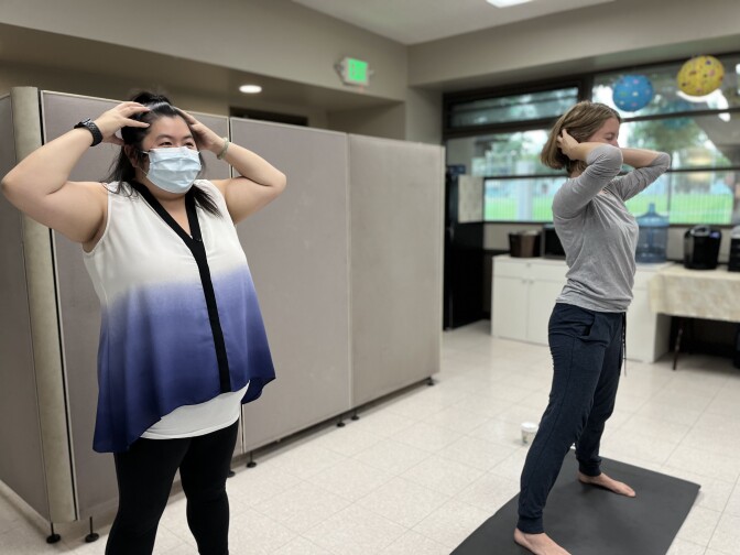 An Asian American woman in a mask and purple and white sleeveless tunic bends her elbow and touches her head, shadowing the movements of a white woman wearing a gray long-sleeved shirt to her left. 