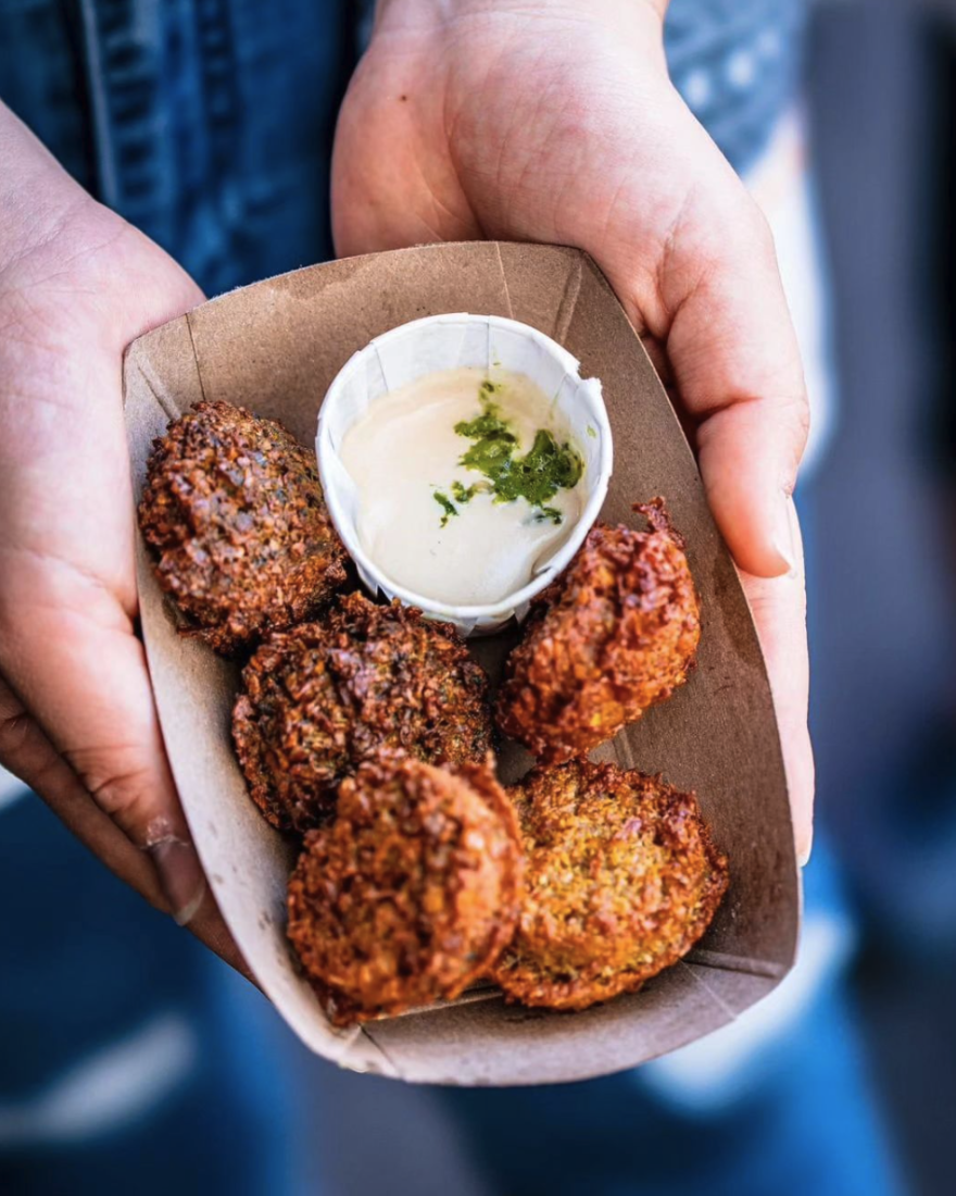 Two hands hold a small cardboard tray containing five falafels that have been so perfectly fried until golden brown that you can practically taste how crunchy they will be. The fritters are clustered around a small paper cup containing a creamy tahini-based dipping sauce topped off with an herb oil.