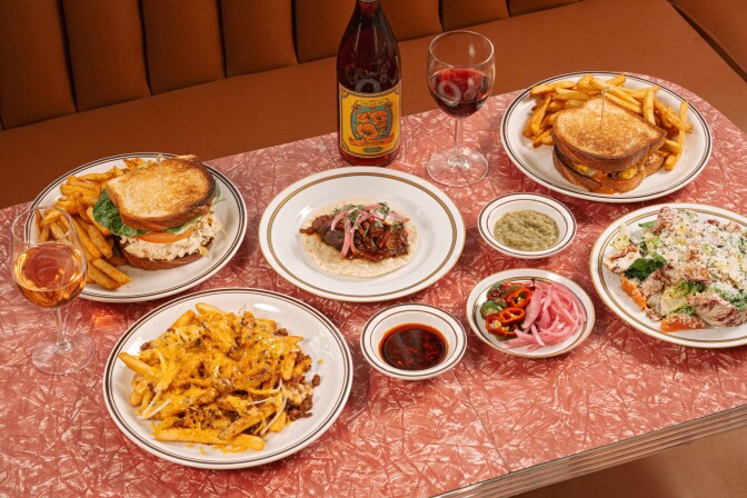 A vibrant, retro-inspired lunch spread set atop a glossy pink cracked-ice Formica table, surrounded by a warm-toned booth featuring  stacked chicken salad sandwich served with golden fries, a single taco , a double-decker patty melt and in the bottom left a plate of loaded chili cheese fries. 