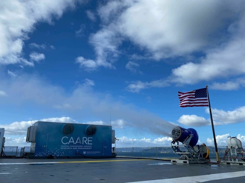 A U.S. flag waves next to a machine expelling material into the air. The sky is cloudy.