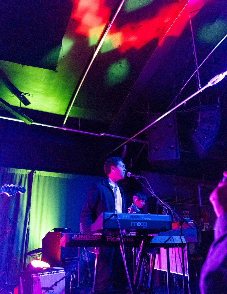 A medium skin tone man with short brown hair and a black jacket stands behind a keyboard and microphone lit by colorful stage lights.