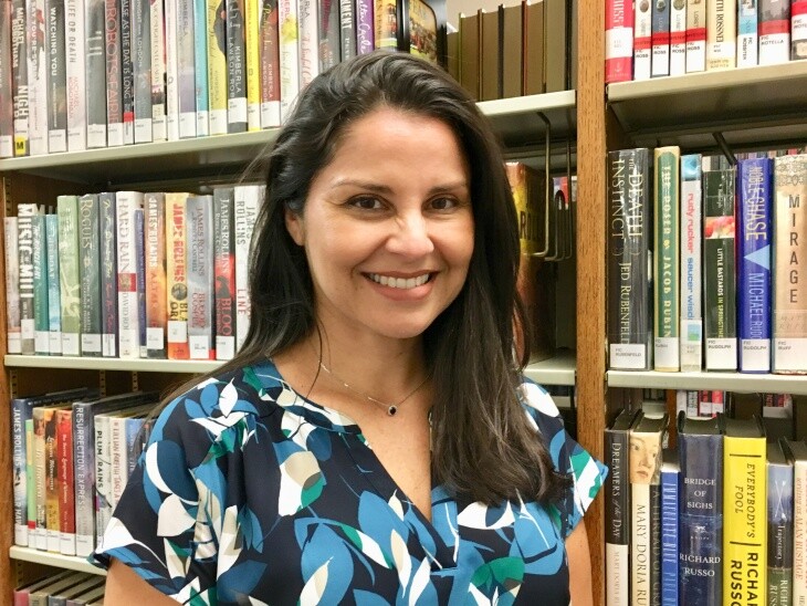A woman with long brown hair stands smiling in front of a bookshelf.