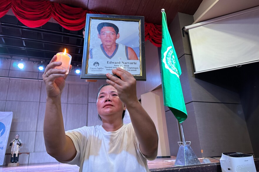 A person with medium-tone skin holds a photo of a man in one hand and a candle in the other.