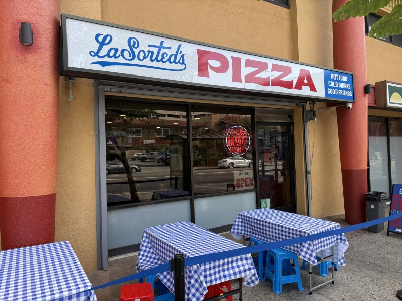 The exterior of LaSorted's pizza restaurant in Chinatown has a sign that reads 'PIZZA' and tables covered with blue and white checked tablecloths.