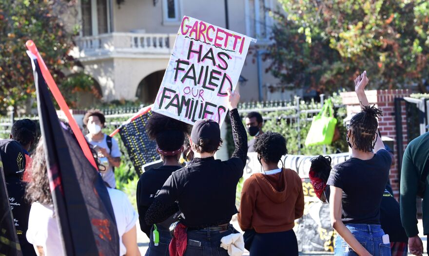 At least a dozen people stand in front of a house. One holds up a sign that reads "Garcetti has failed our families."