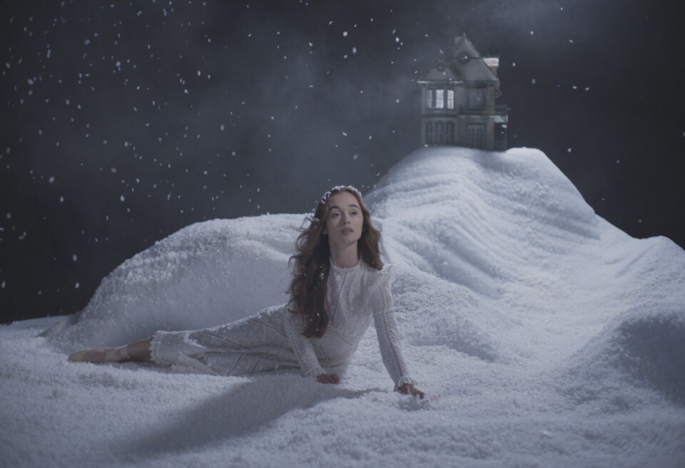 A woman in a white gown lies on a white snow-covered stage scene with a house in the background. 